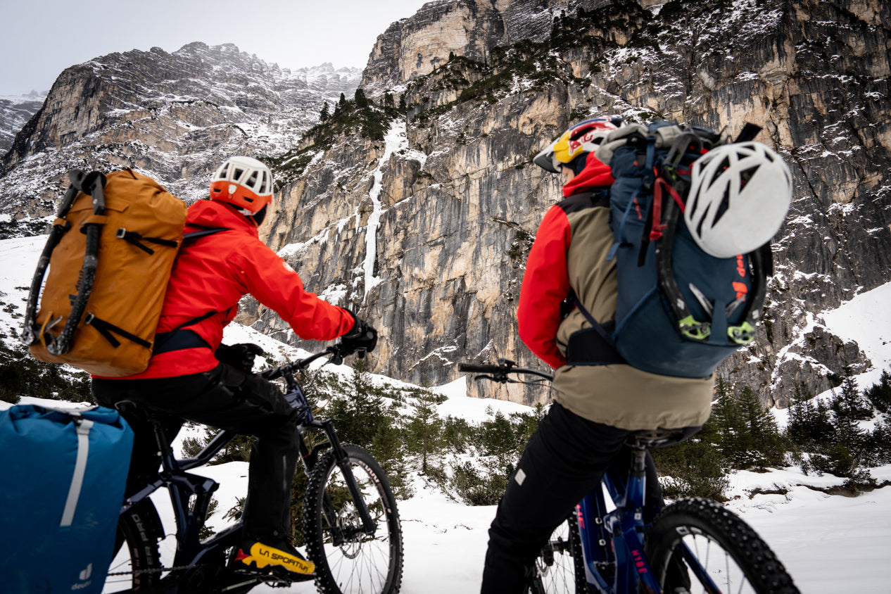 Two mountainbikers with backpacks on Haibike e-bikes with winter camping gear in front of snow-covered rock face on ice climbing adventure tour