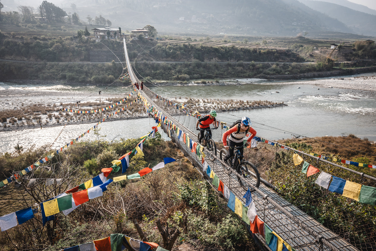 Two Haibike e-mountain bike riders crossing suspension bridge over river in Bhutan lined with colorful Buddhist prayer flags adventure tour 2025