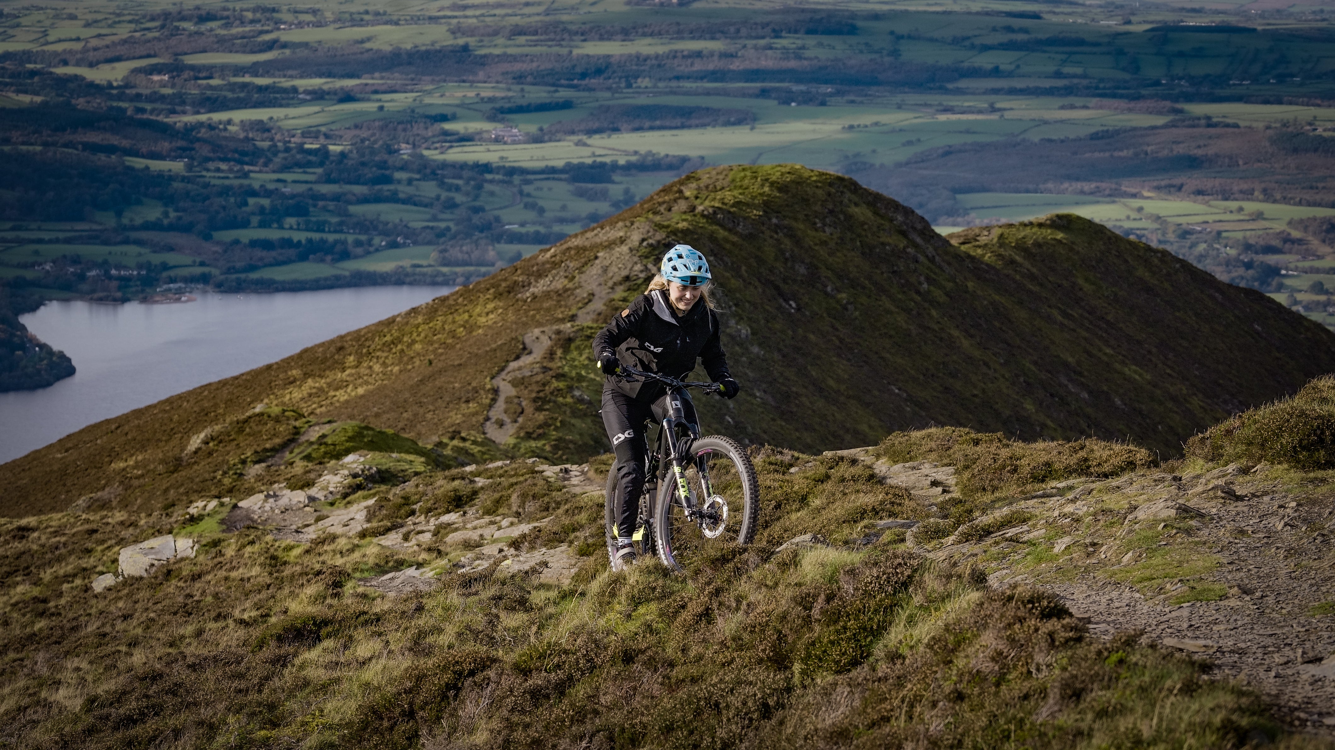 Haibike ambassador Kara Beal riding e-mountain bike down steep highland mountain slope with scenic valley lake backdrop blue helmet e-MTB adventure